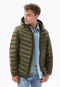 Giubbotto imbottito verde oliva con cappuccio, texture trapuntata, cerniera metallica e tasche laterali. Presenta un marchio rosso sulla manica e una camicia blu sotto.