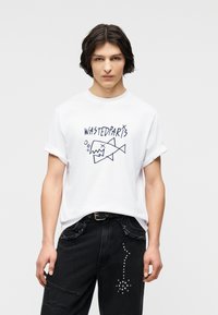 Νεαρός με σκούρα μαλλιά φοράει λευκό μπλουζάκι με γυρισμένα μανίκια που φέρει το κείμενο "WASTEDPARTS" και αφηρημένο γραφικό ψαριού, συνδυασμένο με μαύρο τζιν με διακοσμητικά.