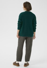 Grüner Strickpullover mit lockerer Passform und gerippten Bündchen, kombiniert mit grauen schmal geschnittenen Hosen und braunen Schuhen, vor einem schlichten Hintergrund.