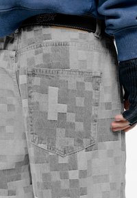 Pantaloni in denim grigio a fantasia con tasca a toppa, caratterizzati da un design pixelato e una texture a contrasto, abbinati a un maglione blu e guanti senza dita.