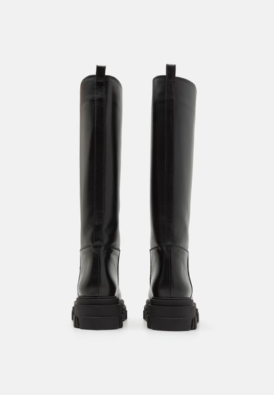 Črne škornje do kolen, izdelane iz gladkega usnja z debelim gumijastim podplatom in vertikalnimi šivi. Dve zanki na vrhu naprave.