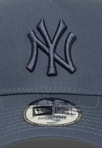 Siva kapa New Era z dvignjenim mornarsko modrim NY logotipom, ki ima nastavljiv trak in okroglo oznako, ki vsebuje informacije o velikosti in blagovni znamki.