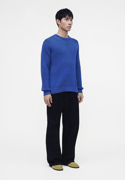 Homme debout portant un pull côtelé bleu, un pantalon noir large et des chaussures jaunes à enfiler sur un fond clair uni.
