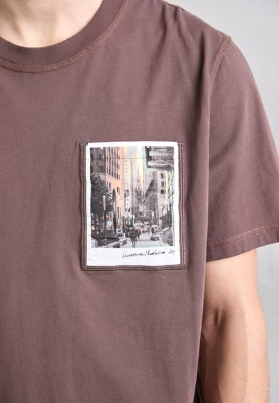 Barna pamut póló rövid ujjal, négyzet alakú zsebfotóval, amely egy városképet ábrázol fekete-fehérben színes kiegészítésekkel.
