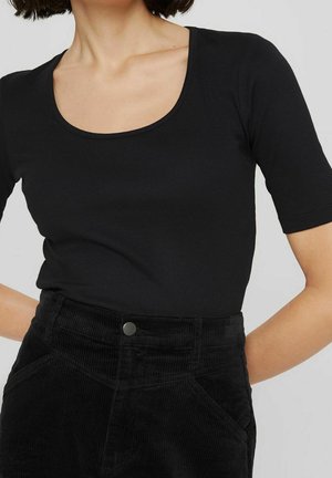 Mujer con una camiseta negra de cuello redondo y mangas cortas, metida dentro de unos pantalones de pana negros de cintura alta con bolsillos delanteros.