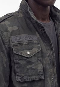 Suurplaan tume kamuflaažvärvi M65 GIANT jakkidest, millel on klapitasku ja tõmblukk, kantud halli särgi peal.