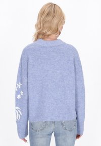 Ljusblå, fluffig stickad tröja med vid passform, ribbat avslut och blommig vit broderi på vänster ärm. Enkel, mjuk textur.