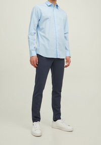 Light blue cotton button-up shirt with long sleeves, paired with dark navy trousers and white sneakers, standing on a neutral background.