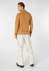 Brun zip-up fleecejakke med høj krave, parret med hvide cargo-snebukser. Outfittet har kontrasterende syninger og sorte accenter.