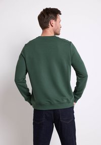 Felpa verde con maniche lunghe e polsini a costina, caratterizzata da un collo rotondo e una texture liscia. Indossata con jeans in denim scuro.
