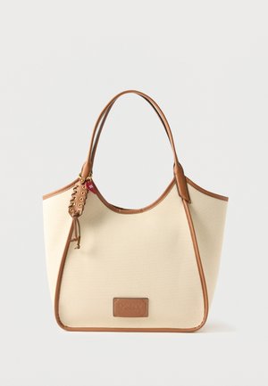 Kremhvid lærredstaske med brune læderkanter, to håndtag, snøredetalje og en lille rød hjertecharm på den ene side.