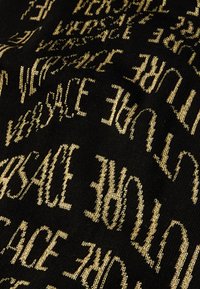 Cachecol tricotado com um fundo preto e texto em dourado "VERSACE" repetido em várias orientações. Superfície texturizada com acabamento suave.