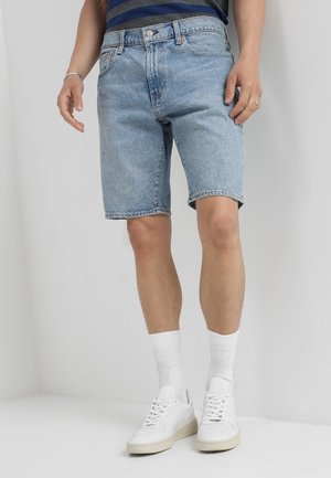 Shorts en denim bleu clair avec une coupe classique, présentant un ourlet effiloché, un design à cinq poches, et portés avec des baskets blanches et des chaussettes.