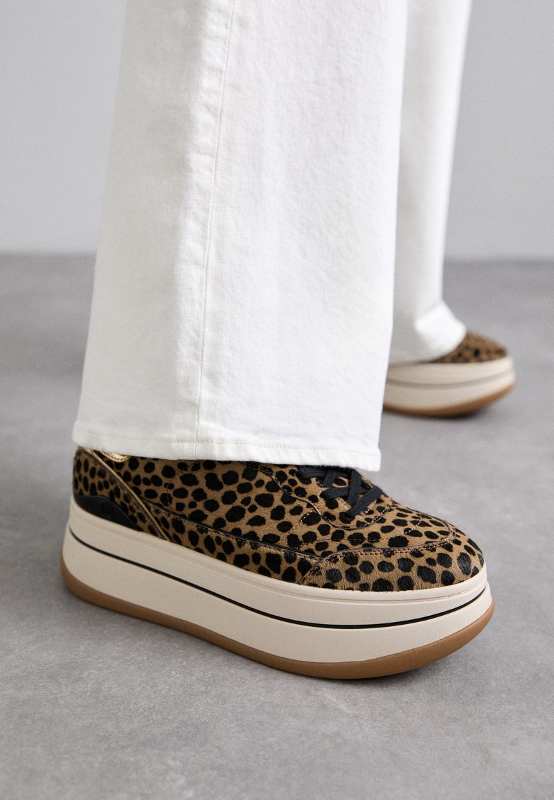 Løveprikkede plattform sneakers med sorte snører, tan- og hvit såle, teksturert overdel og glatte accenter. Brukt sammen med vide hvite bukser.