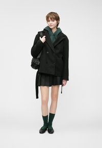 Black double-breasted coat over a green hoodie, paired with a pleated black skirt, green socks, and black shoes, with a small black bag.