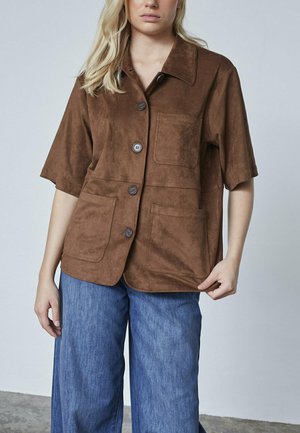 Ung person iført en brun knaplukket kortærmet suedejakke med tre forlommer, parret med løse blå denimjeans.