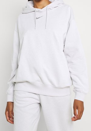 Personne portant un sweat à capuche Nike gris clair et un pantalon de survêtement assorti avec le logo Nike visible sur la poitrine, debout sur un fond blanc.