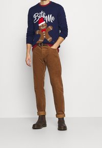 Marineblauer Pullover mit einem Lebkuchenmann-Design und dem Text "Biss mich", kombiniert mit braunen Cordhosen und braunen Chelsea-Stiefeln.