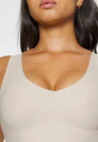 Beige, tettsittende tanktop med V-hals, laget av glatt, elastisk stoff. Kjent for sitt minimalistiske design og sømløse utseende.