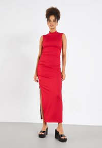 Red ribbed maxi dress with a high collar, featuring side slit and ruched detailing. Model wears black platform heels. Smooth texture.