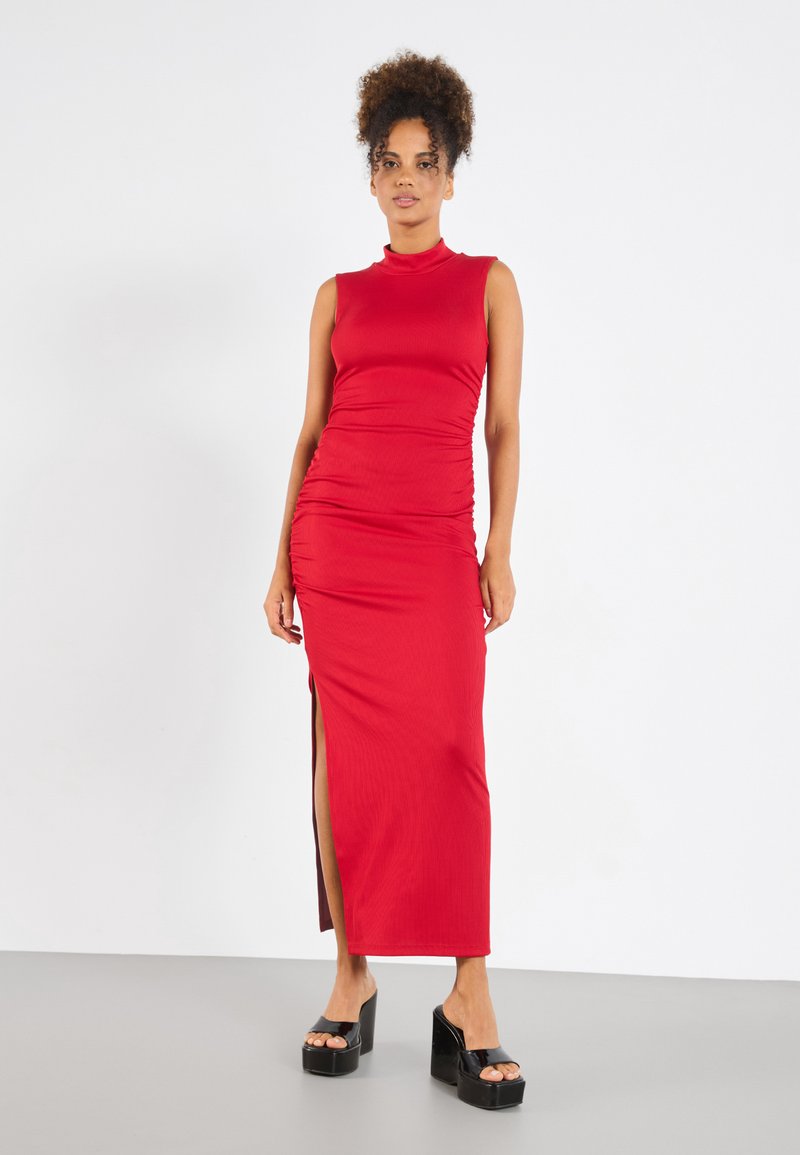 Red ribbed maxi dress with a high collar, featuring side slit and ruched detailing. Model wears black platform heels. Smooth texture.