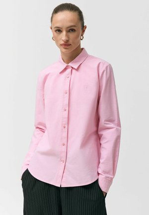 Kobieta z włosami związanymi do tyłu, nosząca złote kolczyki koła, jasnoróżową koszulę na guziki oraz czarne spodnie w prążki, stojąca z rękami w kieszeniach.