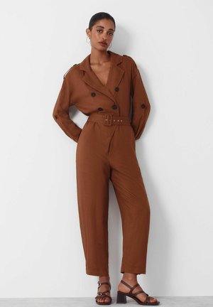 Kvinde står op ad en hvid væg iført en brun jumpsuit med bælte, dobbeltradede knapper og mørkebrune sandaler med blokkappe og stropper.