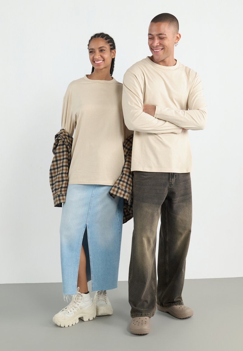Beige long-sleeve shirt on man, paired with faded dark jeans. Woman in beige top, plaid shirt, light denim skirt with slit, white sneakers.