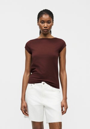 Γυναίκα με πλεγμένα μαλλιά, φορώντας εφαρμοστή κοντομάνικη σκούρα καφέ μπλούζα και λευκά τζιν σορτς με άγρια τελειώματα, στέκεται μπροστά σε απλό φόντο.