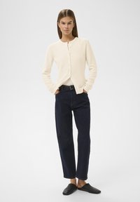 Donna in piedi con le mani nelle tasche, che indossa un cardigan crema a bottoni, jeans scuri a vita alta, cintura nera e scarpe slip-on nere.