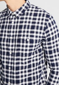 Camicia di flanella a scacchi blu navy e bianca con colletto button-down, maniche lunghe, taschino frontale e bottoni bianchi. Tessuto di cotone, motivo casual.