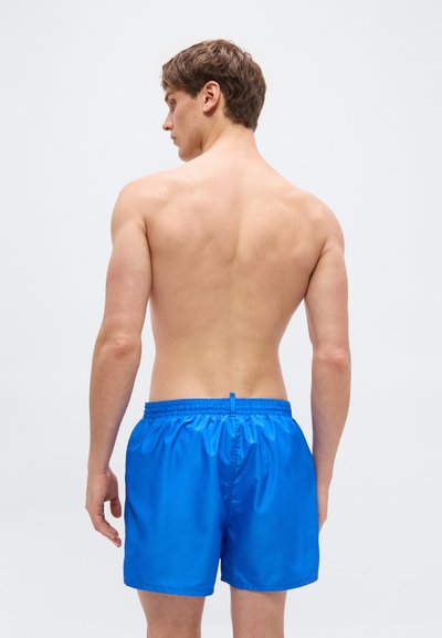 Jeune homme debout, dos à la caméra, portant un short de bain bleu vif sur un fond clair uni.