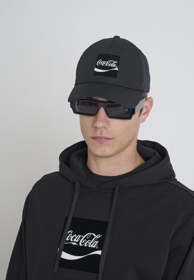 Gorra negra con el logo de Coca-Cola, combinada con una sudadera negra que presenta un logo similar. Modelo usando gafas oscuras. Diseño simple y moderno.