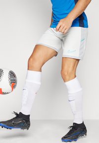 Shorts de football Nike en gris clair avec des accents bleus, associés à des chaussettes blanches et des crampons noirs. Un ballon de football est positionné à proximité.