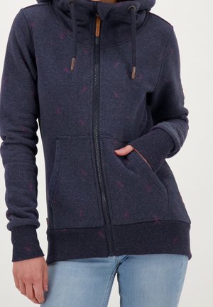 Femme portant un sweat à capuche zippé bleu marine avec des taches roses et des cordons, main dans la poche avant, associée à un jean bleu clair.