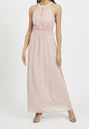 Femme portant une robe maxi sans manches rose pâle avec un détail en dentelle à la taille et une encolure goutte d'eau, associée à des sandales dorées à talons hauts et à brides.