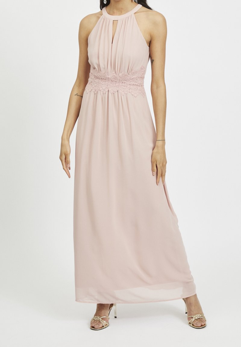 Femme portant une robe maxi sans manches rose pâle avec un détail en dentelle à la taille et une encolure goutte d'eau, associée à des sandales dorées à talons hauts et à brides.