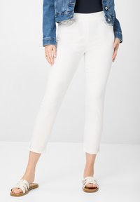 Witte enkellange broek met een elastische tailleband, voorzien van zijzakken en decoratieve uitsparingen aan de onderkant. Gecombineerd met lichte sandalen.
