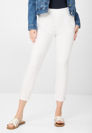 Witte enkellange broek met een elastische tailleband, voorzien van zijzakken en decoratieve uitsparingen aan de onderkant. Gecombineerd met lichte sandalen.