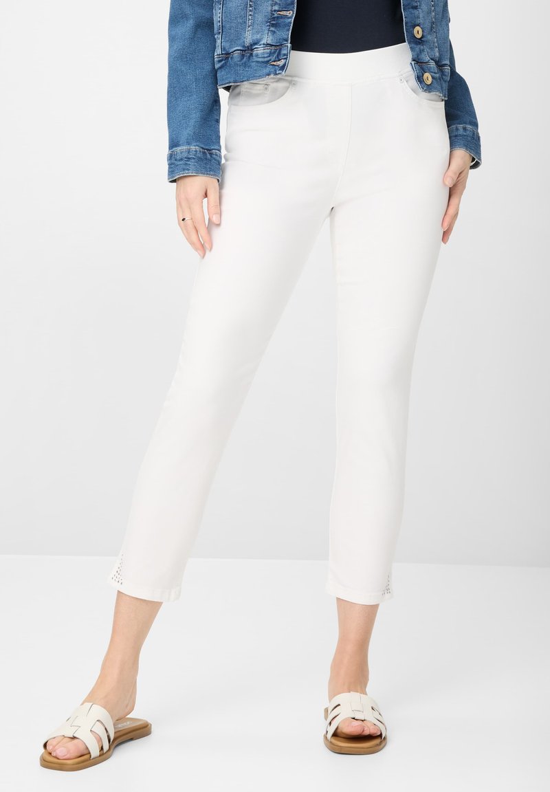 Witte enkellange broek met een elastische tailleband, voorzien van zijzakken en decoratieve uitsparingen aan de onderkant. Gecombineerd met lichte sandalen.