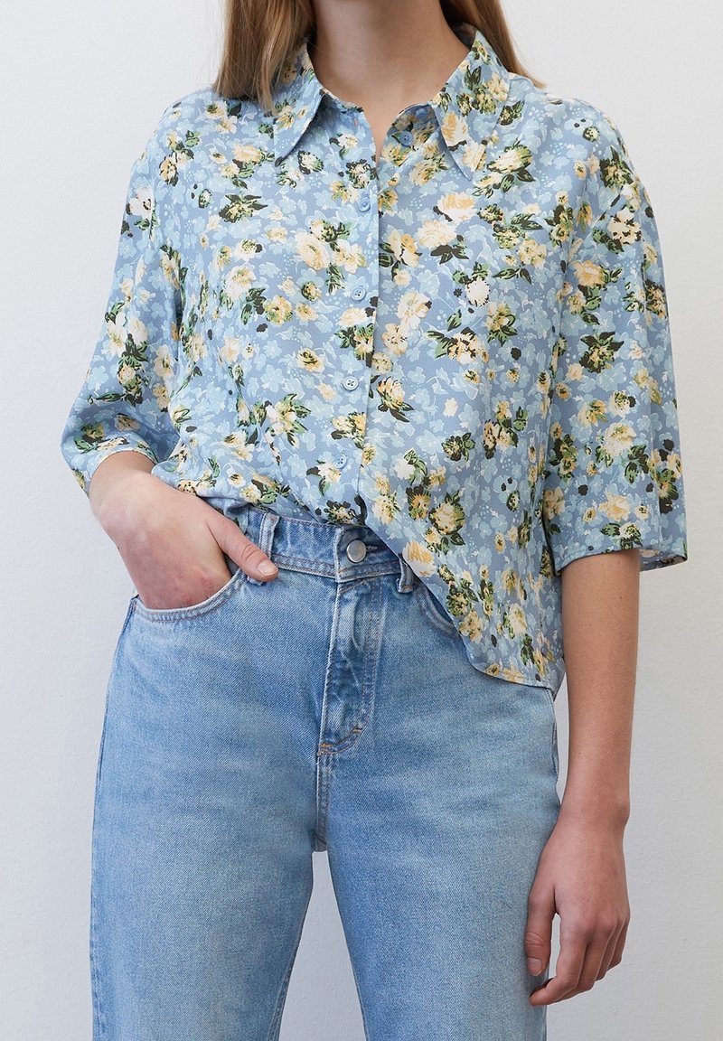 Personne portant une chemise bleu clair à fleurs boutonnée rentrée dans un jean bleu taille haute, avec une main dans la poche.