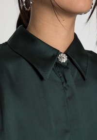 Detailní záběr na límec tmavozelené saténové blůzy, zapnutý dekorativní kulatou broží se rhinestones, kterou nosí osoba s svázanými vlasy.