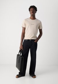 Tricou din bumbac bej cu imprimeu "Arrivederci", blugi din denim bleu închis, geantă din piele neagră și sandale plate negre. O ținută simplă și lejeră.