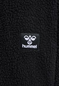 Sort tekstureret stof med et sort rektangulært mærke med en stiliseret hvid bi-logo og ordet "hummel" under.