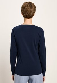 Eine Person mit kurzen braunen Haaren, die ein langärmliges dunkelblaues Hemd und eine hellblaue Hose trägt, steht mit dem Rücken zu einem neutralen Hintergrund.