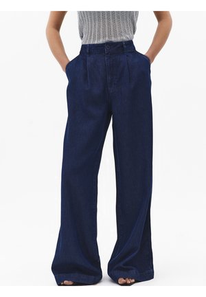 Osoba ubrana w ciemnoniebieskie, szerokie jeansy z wysokim stanem, z rękami w kieszeniach i szary bezrękawnik, stojąca na białym tle.