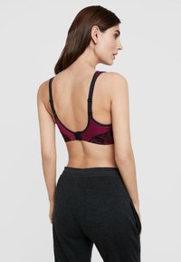 Soutien-gorge de sport bourgogne avec un motif géométrique noir, bretelles ajustables et fermeture à crochets, associé à un pantalon de survêtement gris foncé.