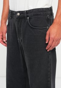 Mand iført sorte denimjeans og en hvid skjorte, hænderne afslappet ned langs siden, viser forlommerne og knaplukningen.