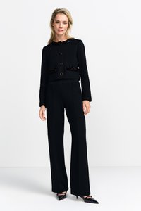 Giacca nera corta con scollo rotondo, dettagli in paillettes sulle tasche e bottoni grandi, abbinata a pantaloni neri a vita alta e tacchi.