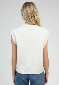 Top en tricot sans manches couleur crème avec un col rond, un ourlet côtelé et des accents aux épaules. Texture légère et douce. Porté avec un jean bleu taille haute.